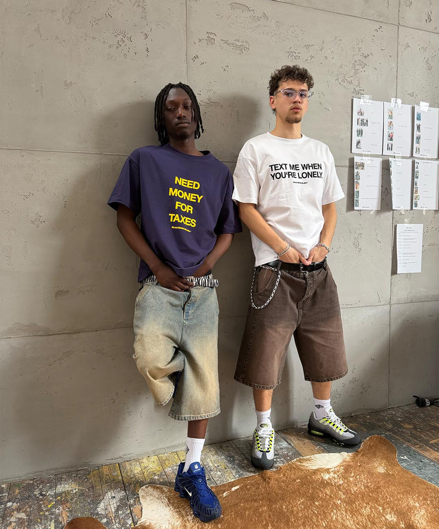 Zwei Freunde lehnen an einer Betonwand, einer trägt das Need Money For Taxes Shirt